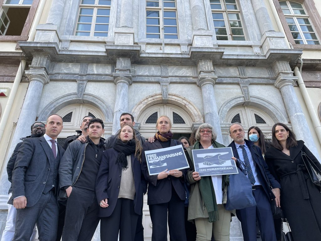 Photo: This morning, Seán Binder stands alongside other defendants and their legal defense team, holding up banners demanding the freedom of relief workers from the steps of the courthouse where the trial is being held on the Greek island of Lesbos. © Posted by Begüm Başdaş on Twitter.